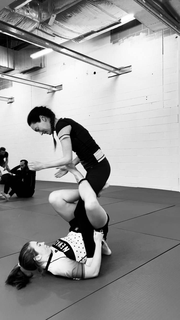 Two students drilling on the mat, one standing in her partner's guard and smiling